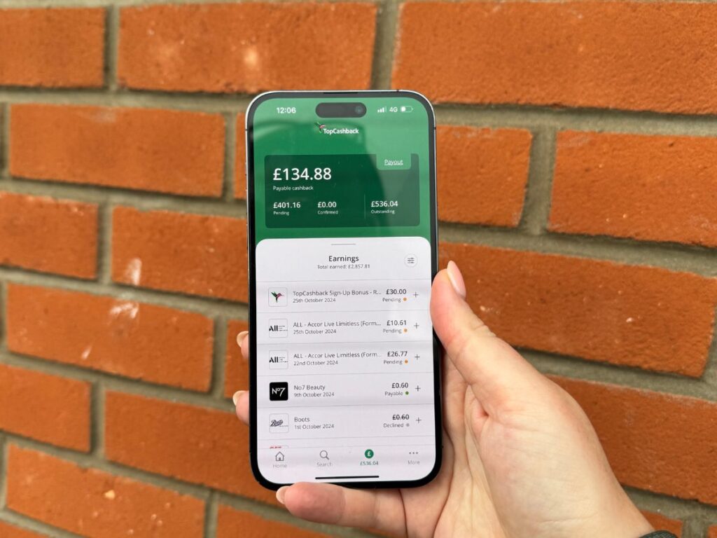 A hand holding a mobile phone, on the screen is the TopCashback app showing earnings on the account