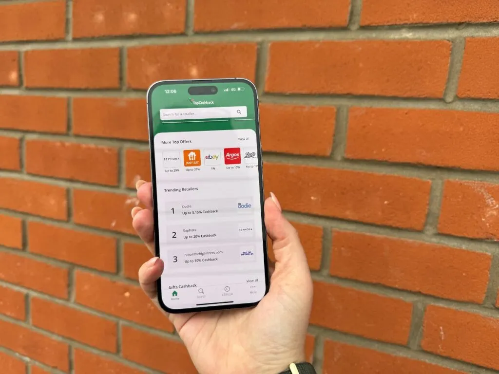 A hand holding a mobile phone displaying the TopCashback app with retailers available and Cashback percentages.