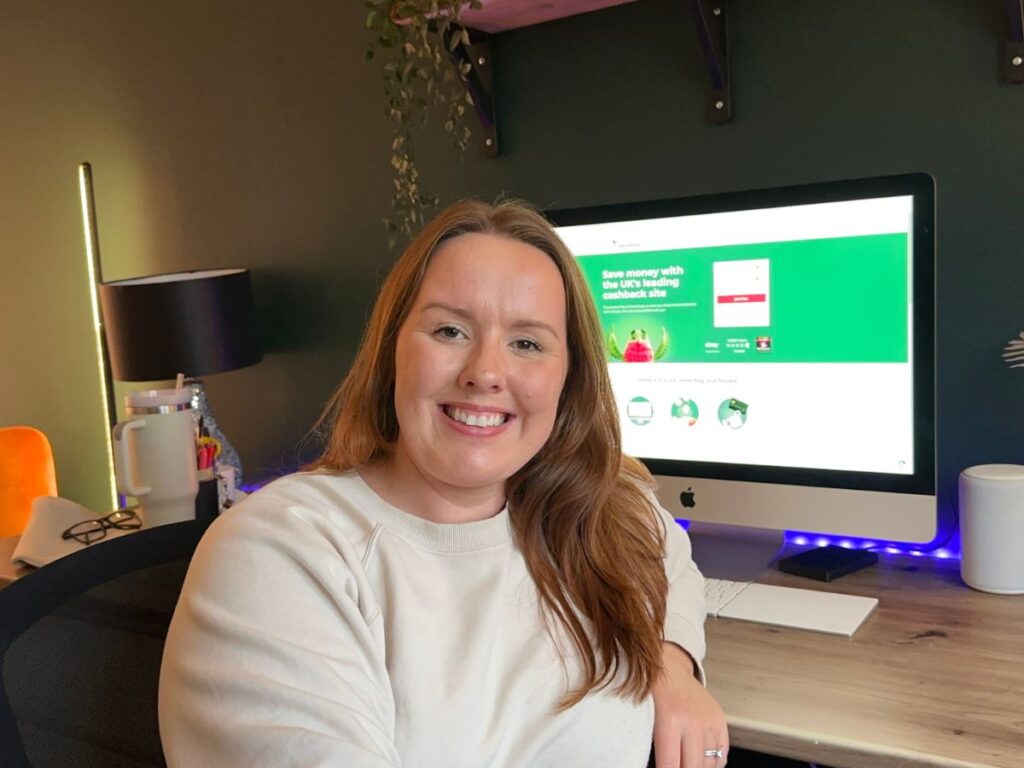 Chloe from Chloe's deal club sitting at a wooden desk with a computer screen in the background, showing the Home Screen for TopCashback