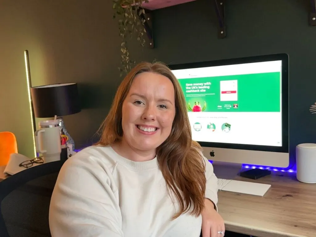 Chloe from Chloe's deal club sitting at a wooden desk with a computer screen in the background, showing the Home Screen for TopCashback