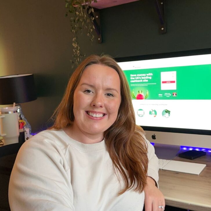 Chloe from Chloe's deal club sitting at a wooden desk with a computer screen in the background, showing the Home Screen for TopCashback