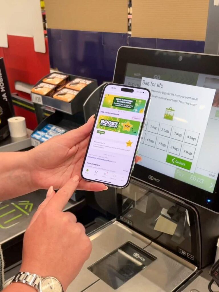 A hand holding a mobile phone with Asda Rewards app on the screen at the Asda checkout.  The screen is showing milestone missions as ways to save money at Asda.