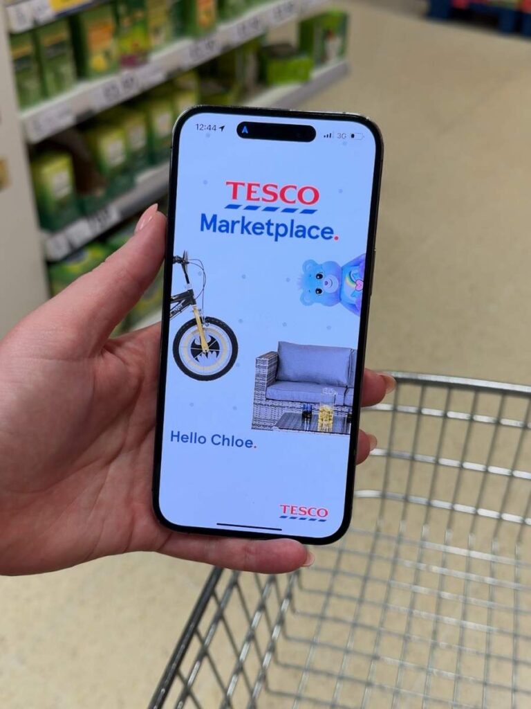 A hand holding a mobile phone displaying the Tesco Clubcard app which shows you how to save money on your shopping